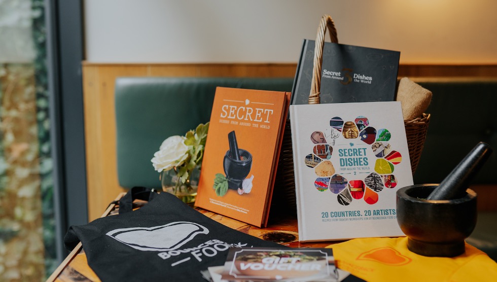 three bounceback food cookbooks propped up on a table, with a tea towel and a apron