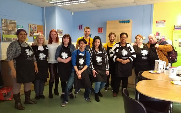 Group of cookery workshops participants and volunteers, wearing BounceBack aprons and stood in a kitchen.