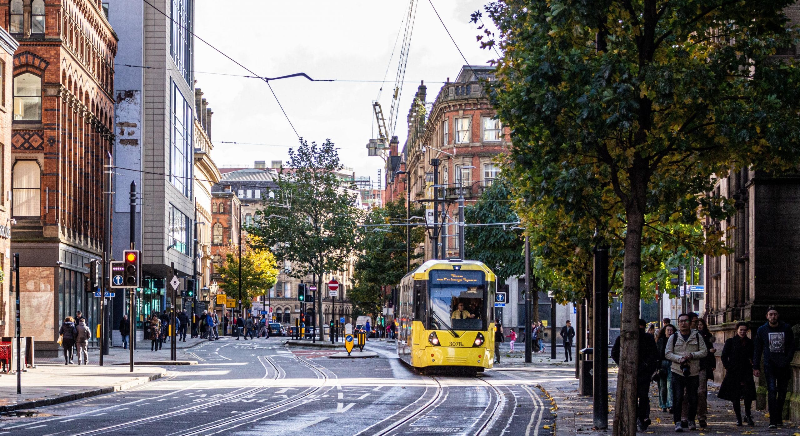 A tram in Manchester city centre on a cloudy day with pedestrians walking down a tree lined street.