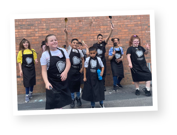 A group of children wearing Bounceback Food Community Cookery School aprons stood outside a school building.