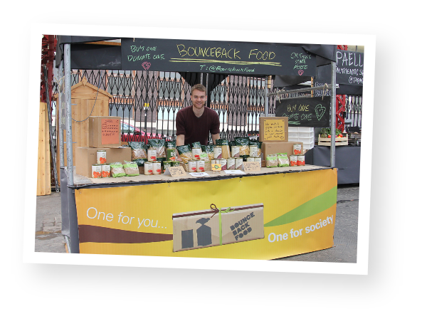 A Bounceback Food employee is stood at a market stall which has a selection of pasta, rice and tinned food for sale.