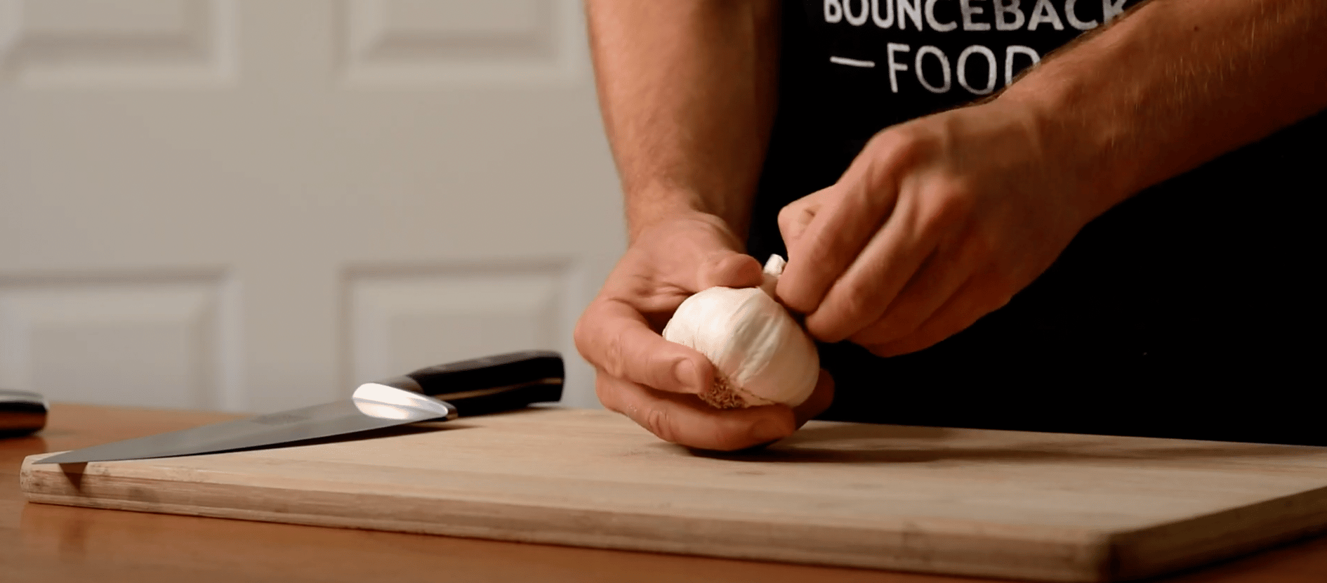 A man in a Bounceback apron is peeling a head of garlic, on a wooden chopping board.