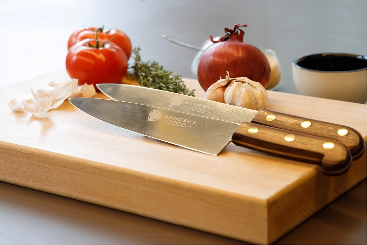 A Bounceback chef's knife placed on top of a chopping board with vegetables in the background.