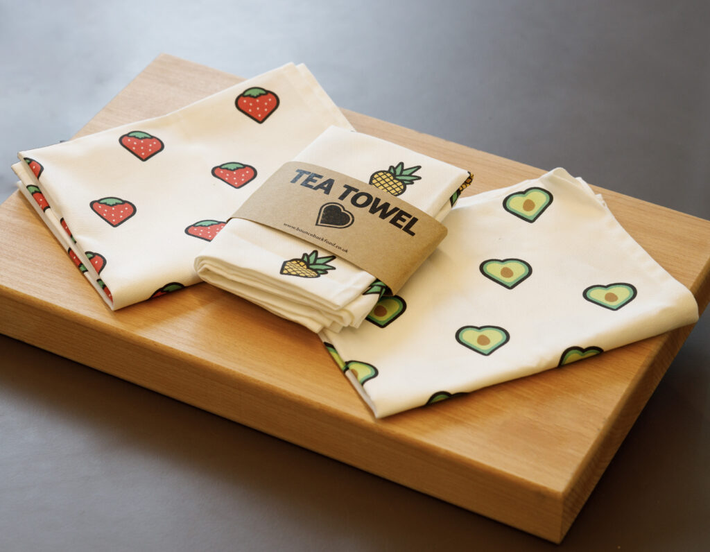 three Bounceback Food tea-towels laid on a wooden chopping board, one with strawberries on, one with pineapples and one with avocado